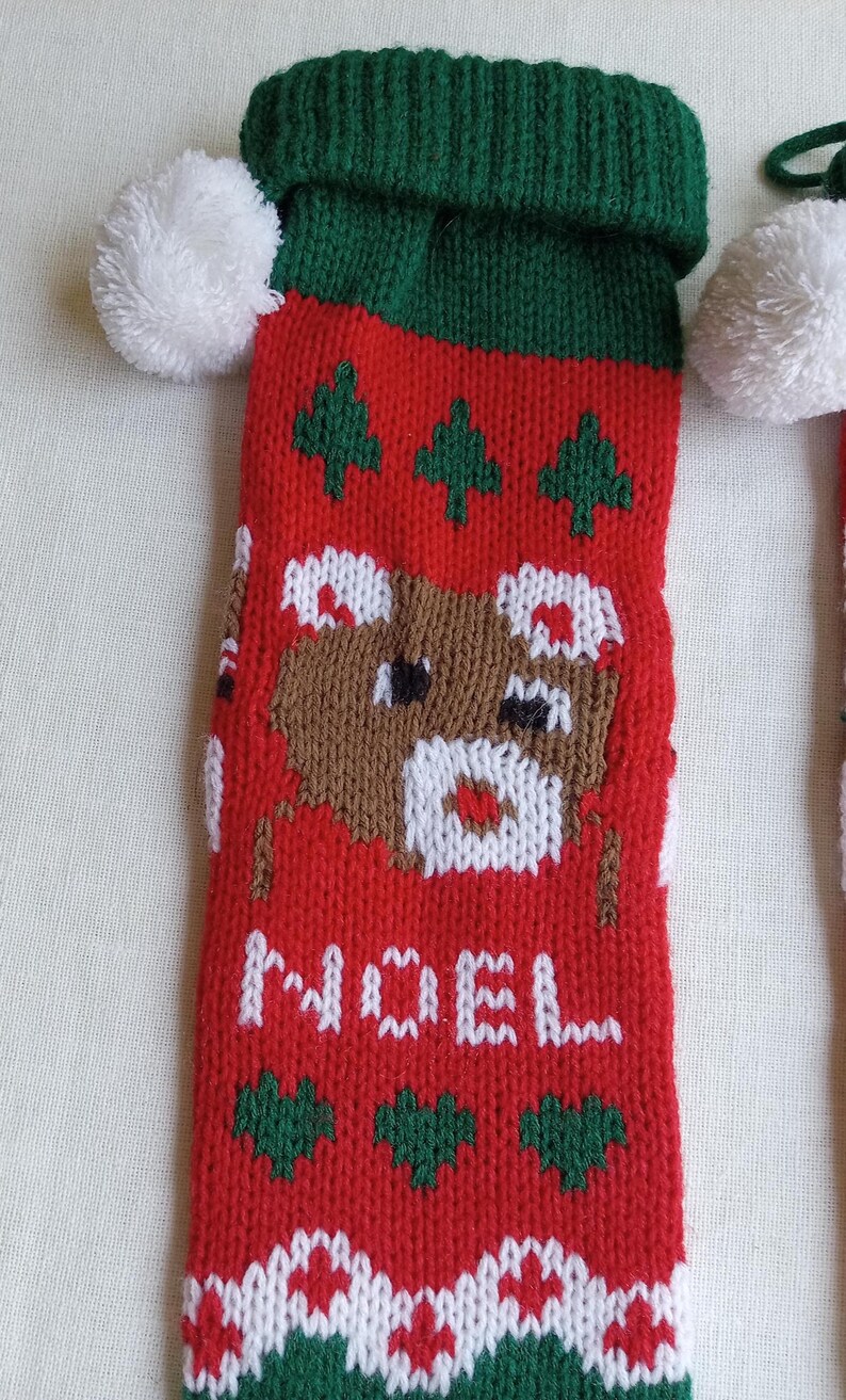 May include: Red Christmas stocking with a green cuff and white pom-pom. The stocking features a knitted design with a brown bear, green trees, and the word "NOEL" in white. A festive holiday decoration.