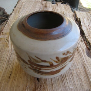 May include: A small, white ceramic pot with a brown rim and brown floral designs. The pot is sitting on a wooden surface.