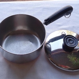 May include: Stainless steel saucepan with a black handle and a matching lid. The lid has a black knob. The pan is round and has a shiny, reflective surface. The handle has a loop for hanging.
