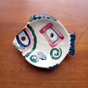May include: A white ceramic fish-shaped dish with a colorful painted design. The design features swirls, lines, and geometric shapes in shades of pink, blue, green, and black.