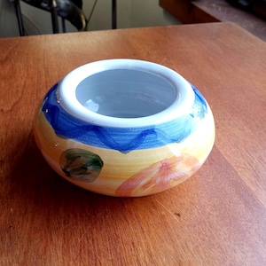 May include: A hand-painted ceramic bowl with a wide opening. The bowl features a design with blue, yellow, and orange colors. The interior is a light gray color. The bowl is sitting on a wooden surface.