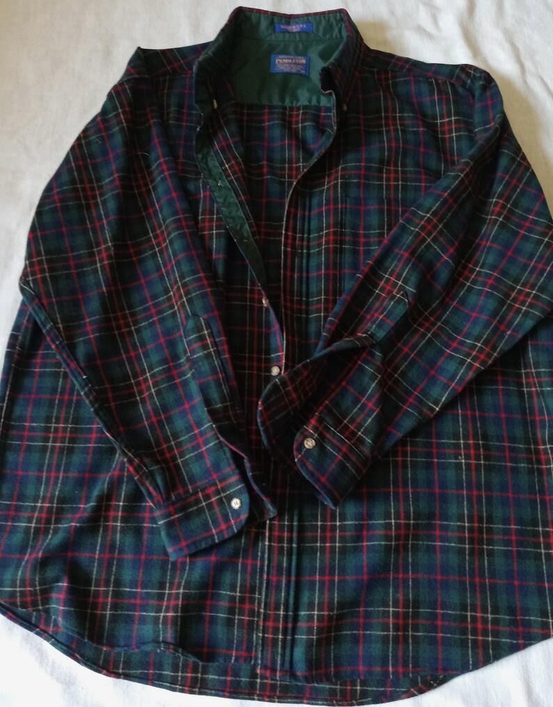 May include: A long-sleeve button-down shirt featuring a dark green, navy blue, and red plaid pattern. The shirt has a dark green inner collar lining, a buttoned front placket, and buttoned cuffs. A Pendleton brand label is visible at the neck.