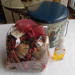 May include: A clear plastic bag filled with potpourri, including cinnamon sticks, pine cones, dried leaves, and berries. The bag is tied with a red ribbon and a tag that reads "Autumn's Perfume". A small clear glass bottle with a spray nozzle contains a liquid. The bottle has a label that reads "Autumn's Perfume". A blue and white tin with a handle is in the background.