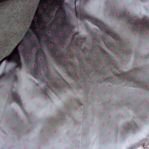 May include: Close-up of the inside lining of a dark gray coat. The lining is a light gray fabric with a repeating pattern of small, pink floral designs. The coat's outer fabric is visible on the left side.