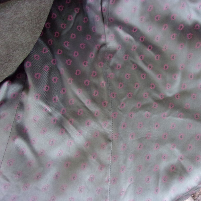 May include: Close-up of the inside lining of a dark gray coat. The lining is a light gray fabric with a repeating pattern of small, pink floral designs. The coat's outer fabric is visible on the left side.