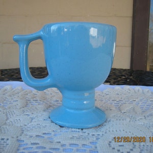 May include: A light blue ceramic mug with a unique handle design. The mug has a pedestal base and a rounded bowl shape. The handle is a curved, rectangular shape. The mug is approximately 4 inches tall.