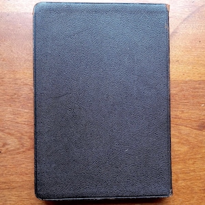 May include: A rectangular book with a dark black, textured cover. The surface of the cover has a fine, pebbled grain pattern. The edges of the book show slight signs of wear with hints of brown material visible along the spine and corners.