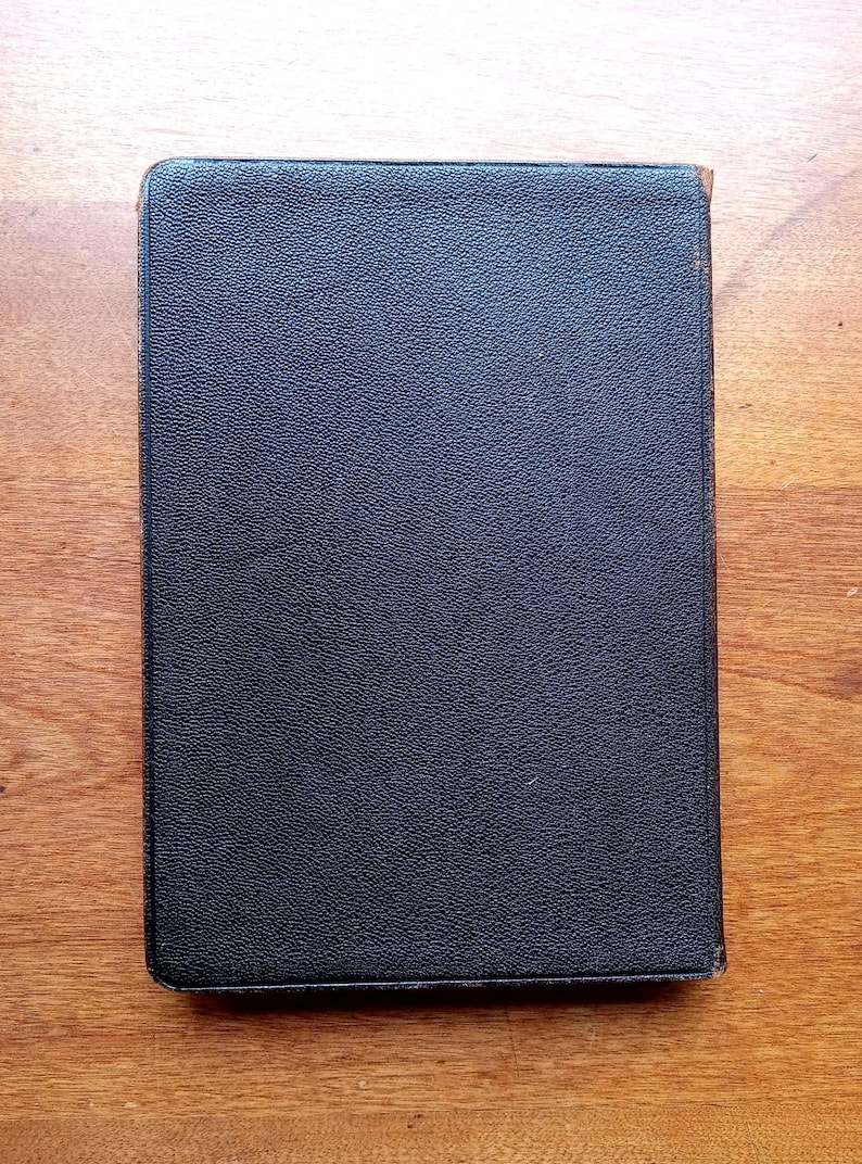 May include: A rectangular book with a dark black, textured cover. The surface of the cover has a fine, pebbled grain pattern. The edges of the book show slight signs of wear with hints of brown material visible along the spine and corners.
