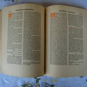 May include: An open book titled Elbert Hubbard's Scrap Book with two pages of printed text. The pages feature black ink columns, decorative orange drop cap letters at the start of paragraphs, and page numbers 134 and 135 visible at the top corners.
