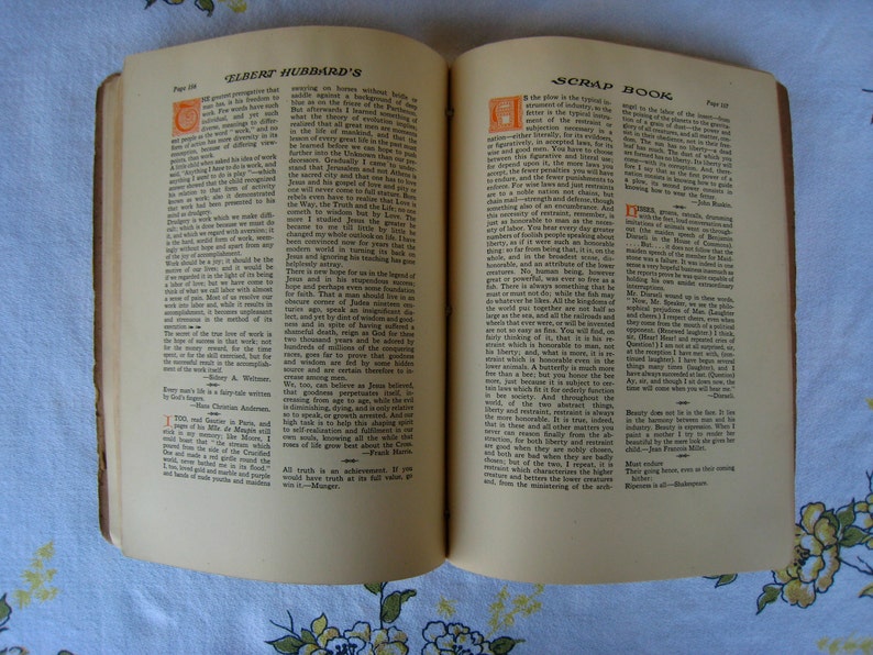 May include: An open book titled Elbert Hubbard's Scrap Book with two pages of printed text. The pages feature black ink columns, decorative orange drop cap letters at the start of paragraphs, and page numbers 134 and 135 visible at the top corners.