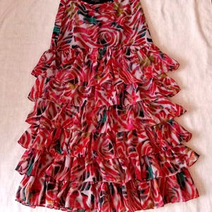 May include: A tiered skirt with a vibrant red, pink, and white abstract floral pattern. The skirt features multiple layers of ruffled fabric, creating a flowing and dynamic silhouette. The design incorporates touches of green and black.