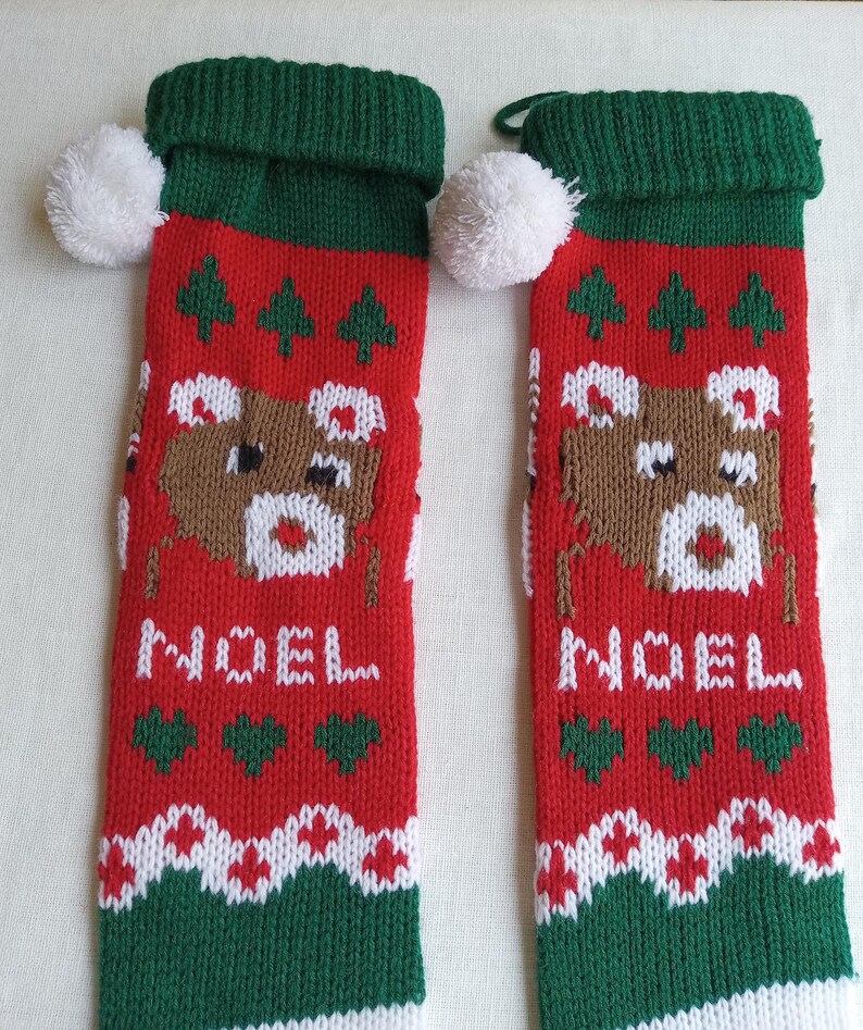 May include: Two red Christmas stockings with green cuffs and white pom-poms. Each stocking features a brown reindeer design, green Christmas trees, and the word "NOEL" in white. White and green patterns decorate the bottom.