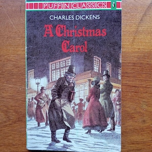 May include: A vintage paperback book titled "A Christmas Carol" by Charles Dickens. The cover features an illustration of people in Victorian-era clothing on a snowy street. The book is part of the Puffin Classics series.