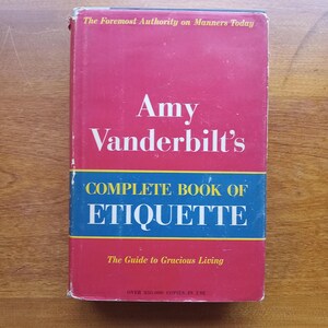 May include: A red hardcover book titled "Amy Vanderbilt's Complete Book of Etiquette: The Guide to Gracious Living". The book cover has gold lettering and a blue band with the word "ETIQUETTE" in white lettering.
