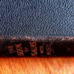 May include: A dark, textured book spine with gold lettering that reads THE BOOK OF PSALMS WITH MUSIC. The cover material appears to be a dark, grained leather or synthetic leather with visible wear along the edges of the spine, showing brown tones.