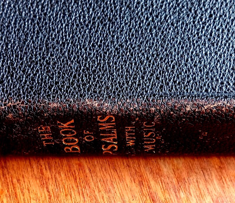 May include: A dark, textured book spine with gold lettering that reads THE BOOK OF PSALMS WITH MUSIC. The cover material appears to be a dark, grained leather or synthetic leather with visible wear along the edges of the spine, showing brown tones.