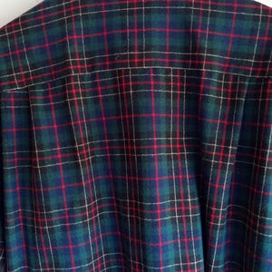 May include: A long-sleeve button-down shirt featuring a classic plaid pattern in shades of dark green, navy blue, and black, accented with thin vertical and horizontal red lines. The shirt has a standard collar and is displayed on a wooden hanger.