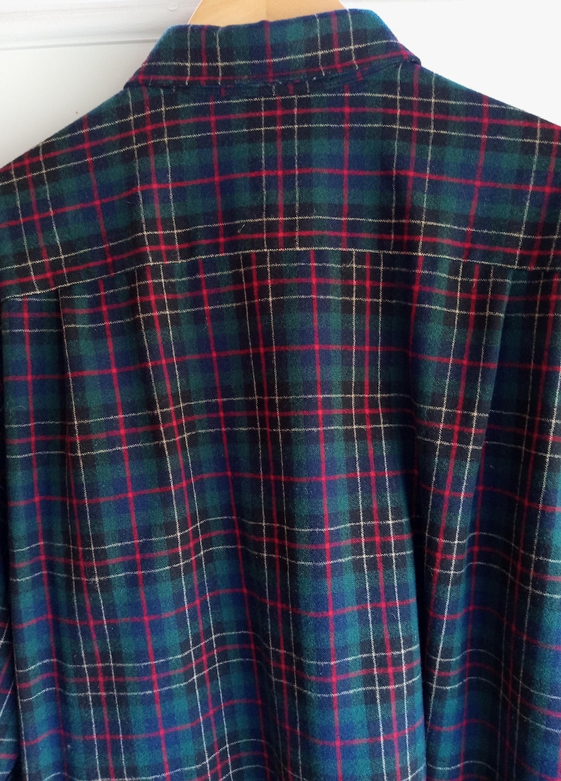 May include: A long-sleeve button-down shirt featuring a classic plaid pattern in shades of dark green, navy blue, and black, accented with thin vertical and horizontal red lines. The shirt has a standard collar and is displayed on a wooden hanger.