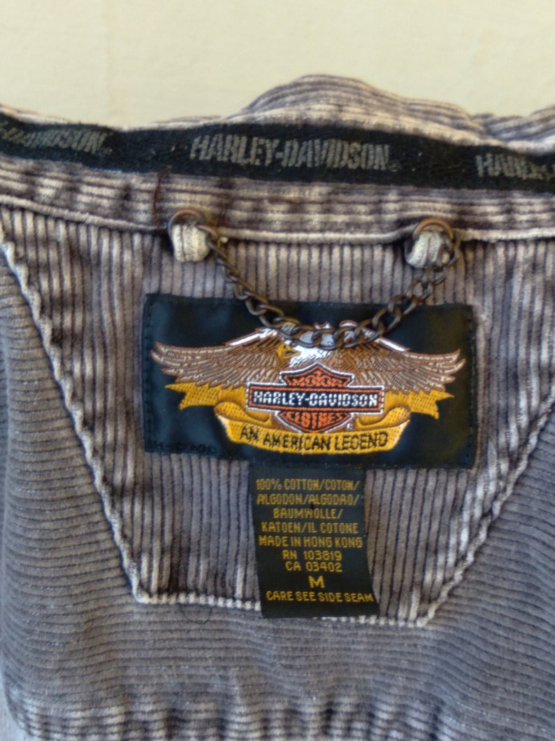 May include: A gray corduroy jacket with a Harley-Davidson logo patch. The patch features an eagle with a shield that says "Harley-Davidson Clothes An American Legend". The tag also says "100% Cotton/Coton/Algodon/Algodao/Baumwolle/Katoen/Il Cotone Made in Hong Kong RN 103819 CA 03402 M Care See Side Seam".
