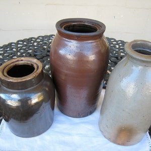 May include: Three vintage stoneware crocks, one brown, one dark brown, and one light gray. The crocks are sitting on a black metal table with a white tablecloth.