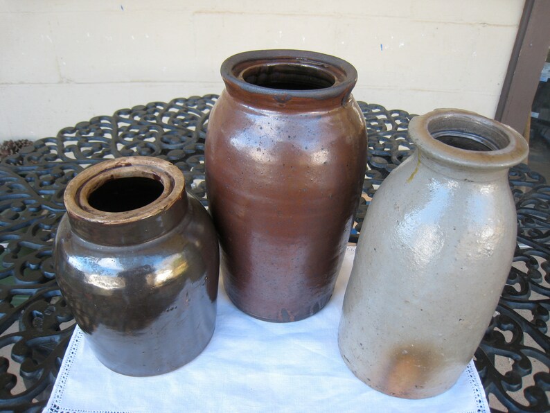 May include: Three vintage stoneware crocks, one brown, one dark brown, and one light gray. The crocks are sitting on a black metal table with a white tablecloth.