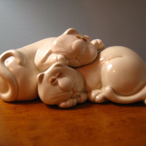 May include: Two white ceramic cat figurines, one lying on top of the other, with their eyes closed. The cats are in a sleeping position, with their tails curled around their bodies.