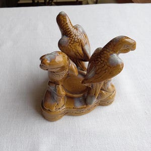 May include: A ceramic sculpture featuring three birds in shades of brown and tan. The birds are perched on a decorative base with intricate details. The sculpture has a glossy finish and is likely a decorative item.