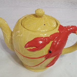 May include: A yellow ceramic teapot with a lobster design. The teapot has a lid and a handle. The lobster is red and has a black eye.