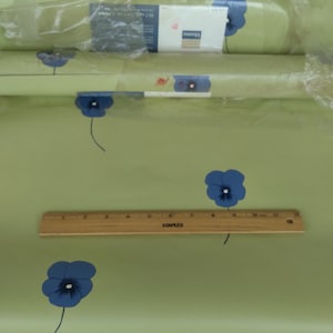May include: A roll of green wallpaper with a blue floral pattern. The wallpaper is packaged in a clear plastic bag and includes a ruler for measuring.