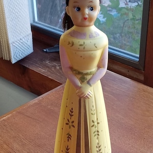 May include: A vintage yellow plastic doll bottle with a brown belt and floral design. The doll has brown hair, blue eyes, and a pink dress.