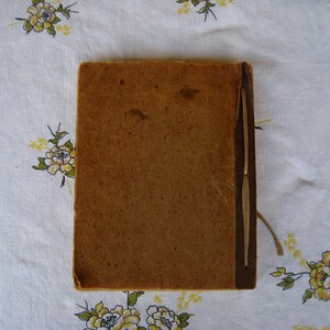 May include: A rectangular book with a weathered, tan-colored cover and a darker brown spine. The cover shows signs of age with visible surface texture, minor discoloration, and wear along the edges. A thin, light-colored string is visible.