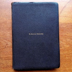 May include: A rectangular, dark navy blue book with a textured, leather-like cover. The name R. Geneva Alexander is embossed in small, gold-colored capital letters in the center of the front cover. The edges of the book show signs of wear.