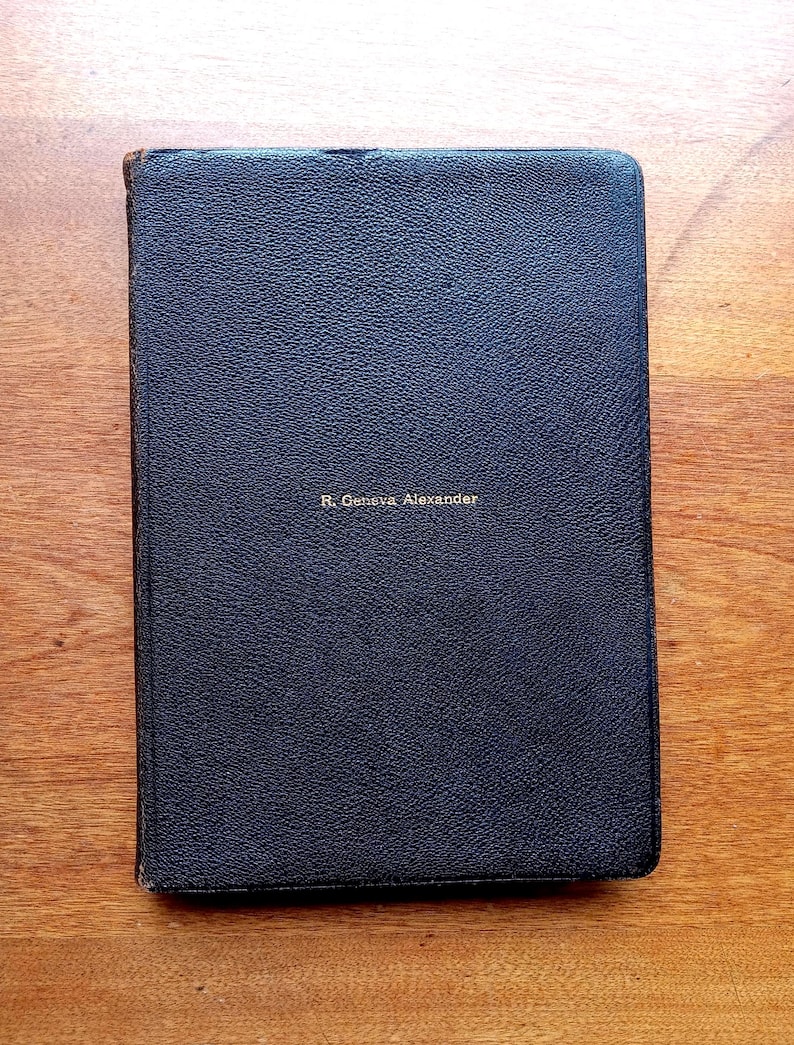 May include: A rectangular, dark navy blue book with a textured, leather-like cover. The name R. Geneva Alexander is embossed in small, gold-colored capital letters in the center of the front cover. The edges of the book show signs of wear.