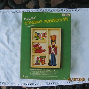 May include: A Bucilla "Toyland" creative needlecraft kit. The green box features a shadow box design with a kite, teddy bear, toy soldier, and train. The kit includes a 5 x 7 inch hutch, yarn, floss, felt, beads, and instructions.
