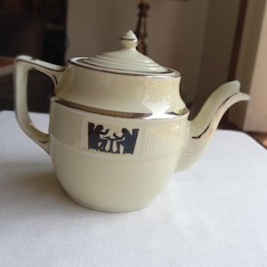 May include: Cream-colored ceramic teapot with a lid and handle, accented with silver trim. The teapot features a black silhouette design of two people at a table. The spout and handle are curved.