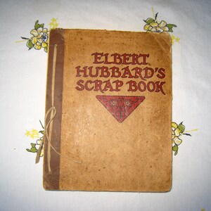 May include: A tan-colored scrap book titled ELBERT HUBBARD'S SCRAP BOOK in red lettering. The cover features a dark brown spine with a tied string binding and a red triangular decorative element below the title with intricate black line details.