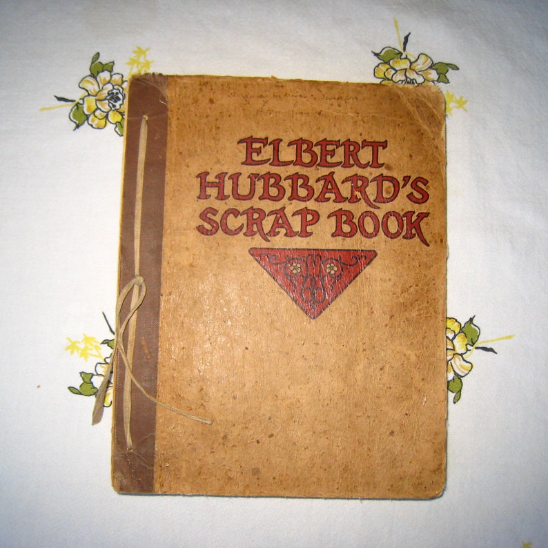 May include: A tan-colored scrap book titled ELBERT HUBBARD'S SCRAP BOOK in red lettering. The cover features a dark brown spine with a tied string binding and a red triangular decorative element below the title with intricate black line details.