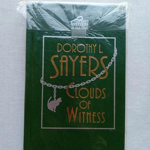 May include: A hardcover book titled "Clouds of Witness" by Dorothy L. Sayers, wrapped in plastic. The cover is dark green with gold lettering and a cat pendant. The text "The Best Mysteries of All Time" is at the top.