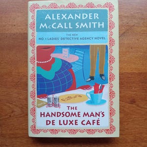 May include: A book cover featuring the title "The Handsome Man's De Luxe Café" by Alexander McCall Smith. The cover art depicts a cafe scene with a woman, a cup of coffee, and the text "The New No. 1 Ladies' Detective Agency Novel."