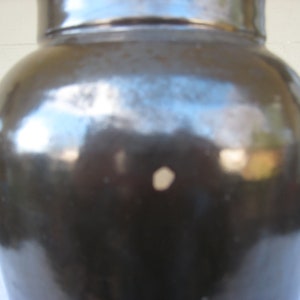 May include: A dark brown ceramic crock with a smooth, glossy finish. The crock has a slight indentation on the side.
