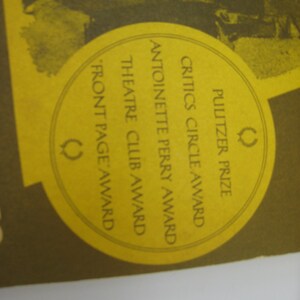 May include: A yellow circle with the text "Pulitzer Prize", "Critics Circle Award", "Antoinette Perry Award", "Theatre Club Award", and "Front Page Award" in black text. A laurel wreath symbol is on the right side of the circle.