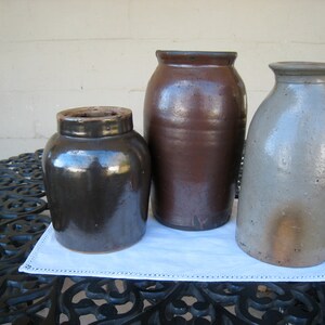 May include: Three vintage stoneware crocks, one brown, one dark brown, and one light gray, sitting on a black metal table with a white doily.