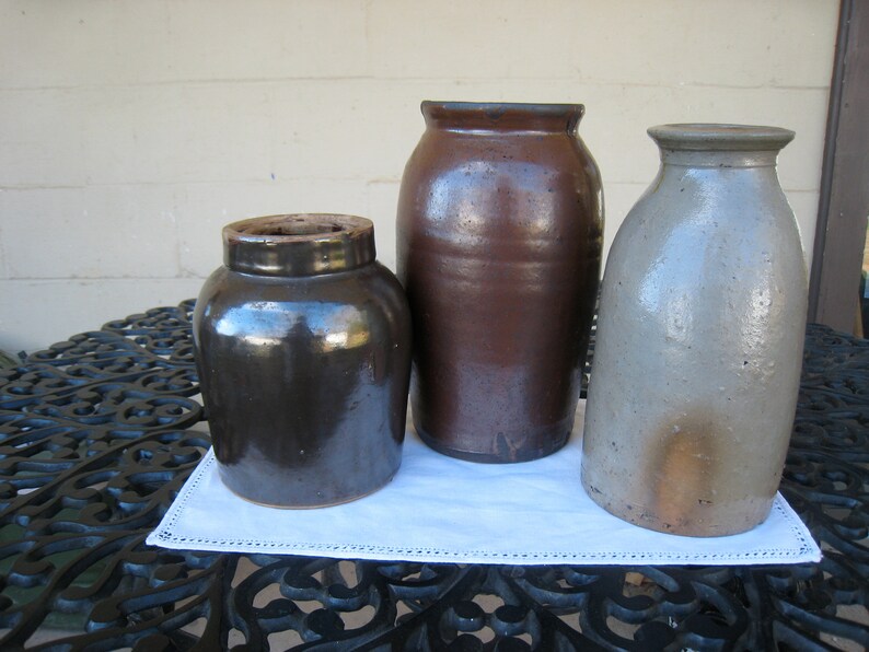 May include: Three vintage stoneware crocks, one brown, one dark brown, and one light gray, sitting on a black metal table with a white doily.