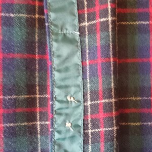 May include: A fabric garment featuring a dark green, navy blue, and red plaid tartan pattern. A vertical strip of solid dark green satin-like material runs down the center, secured with two small, light-colored thread stitches visible on the surface.