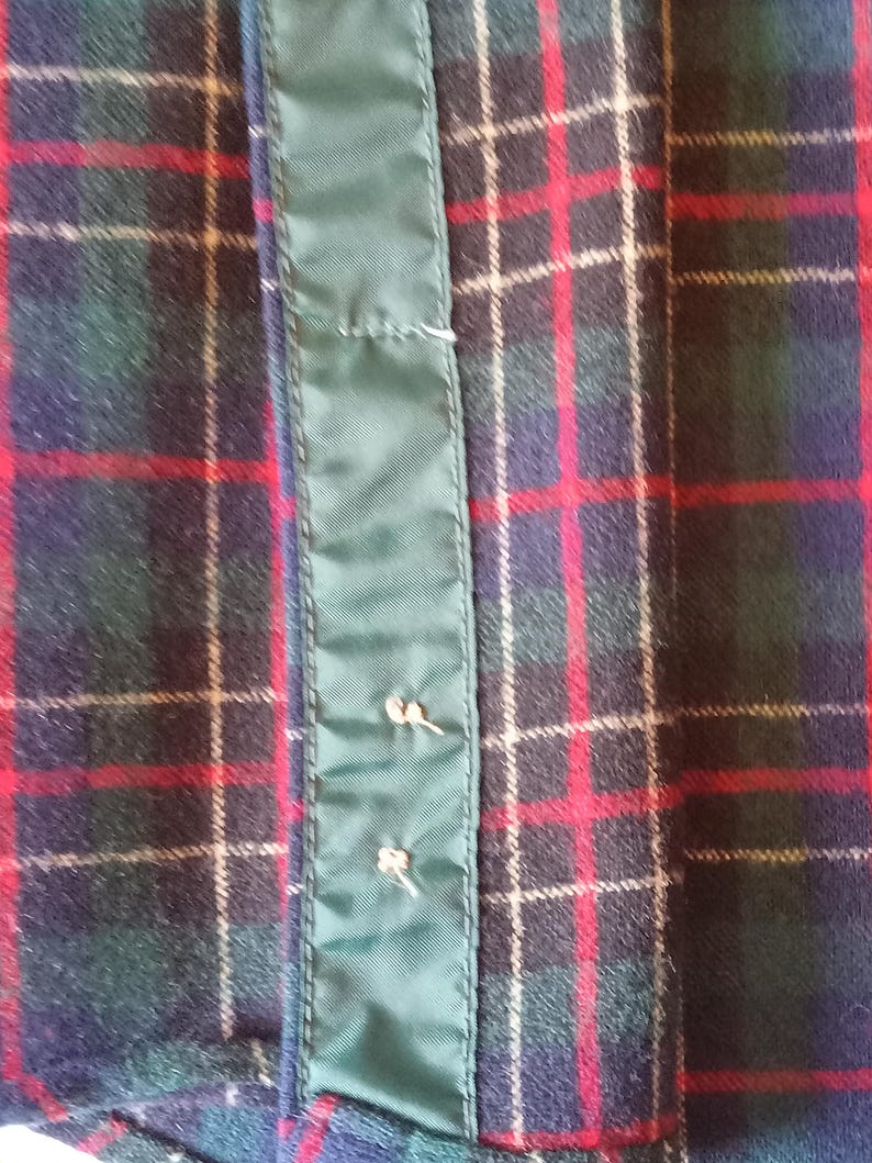 May include: A fabric garment featuring a dark green, navy blue, and red plaid tartan pattern. A vertical strip of solid dark green satin-like material runs down the center, secured with two small, light-colored thread stitches visible on the surface.