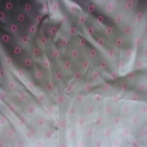 May include: Close-up of a fabric with a repeating pattern of pink floral shapes on a gray background. The fabric appears to be smooth and possibly satin or silk, with visible folds and a seam.