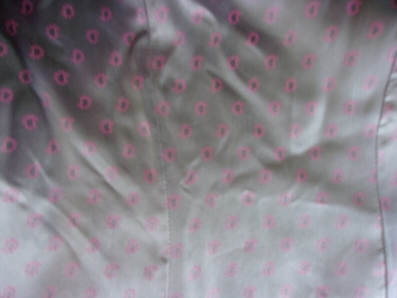 May include: Close-up of a fabric with a repeating pattern of pink floral shapes on a gray background. The fabric appears to be smooth and possibly satin or silk, with visible folds and a seam.
