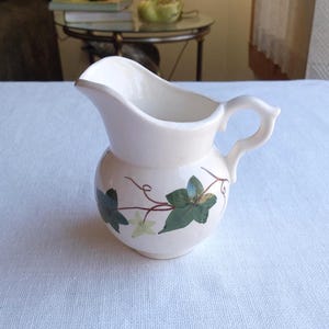 May include: A small, off-white ceramic pitcher with a rounded body and a curved handle. The pitcher features a decorative design of green ivy leaves and vines. The spout is gently curved for easy pouring. A charming addition to any kitchen or dining setting.