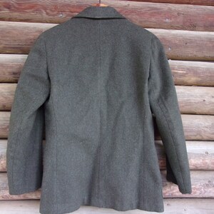 May include: A dark gray wool blazer with a notched lapel and two-button closure. The back of the jacket is visible, hanging on a wooden hanger against a rustic wood background. The blazer has a classic, tailored design.