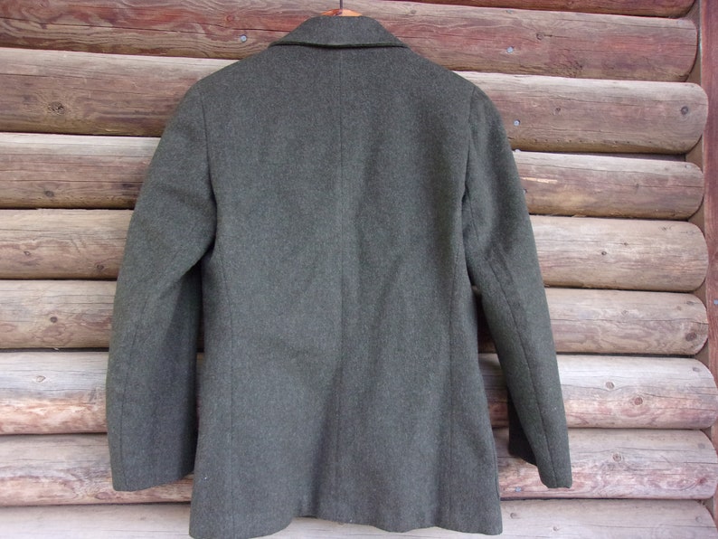 May include: A dark gray wool blazer with a notched lapel and two-button closure. The back of the jacket is visible, hanging on a wooden hanger against a rustic wood background. The blazer has a classic, tailored design.